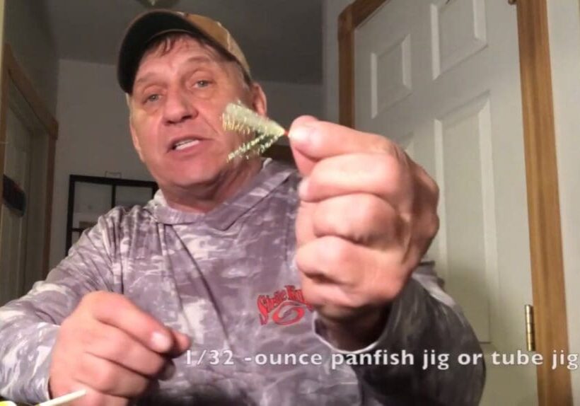 A man in a camo shirt and cap, possibly Mike Frisch, holds up a small fishing jig toward the camera. Text at the bottom reads: "1/32-ounce panfish jig or tube jig.