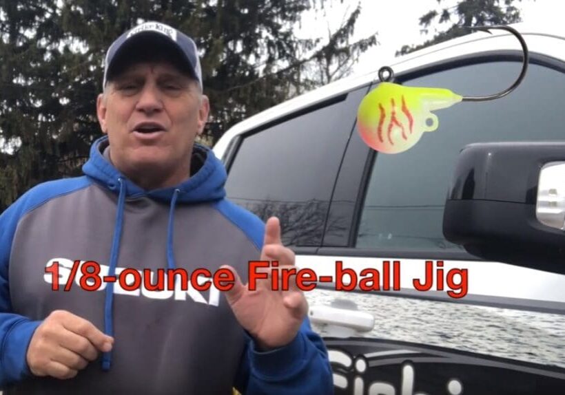 A man in a blue and gray hoodie stands next to a truck, holding up his finger. Overlaid text reads "1/8-ounce Fire-ball Jig" with an image of a yellow and red fishing jig above the text, featuring pro angler Mike Frisch.
