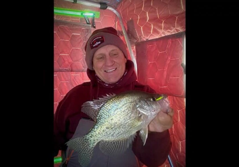 Mike Frisch, in a maroon hoodie and beanie, smiles while holding a large fish inside an ice fishing shelter with red, hexagonal-patterned walls.