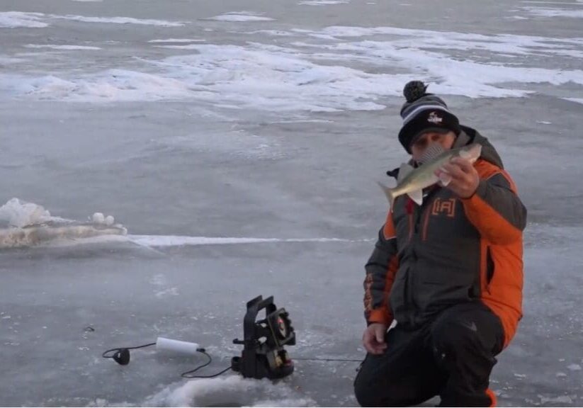 A person in winter clothing kneels on a frozen lake, holding a fish. Ice fishing equipment and a hole in the ice are nearby, surrounded by snow and ice patches—channeling the expertise of mike frisch.