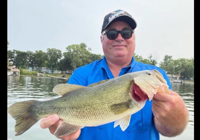 A man in a blue shirt and sunglasses smiles while holding a large summer bass with both hands, standing on a boat with water and trees in the background.