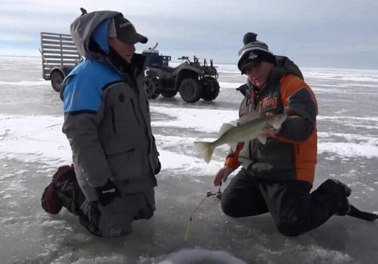 Two people in winter gear kneel on a frozen lake; one, possibly Mike Frisch, holds a fish and a fishing rod. An ATV and sled are in the background, with ice fishing holes visible on the icy surface.