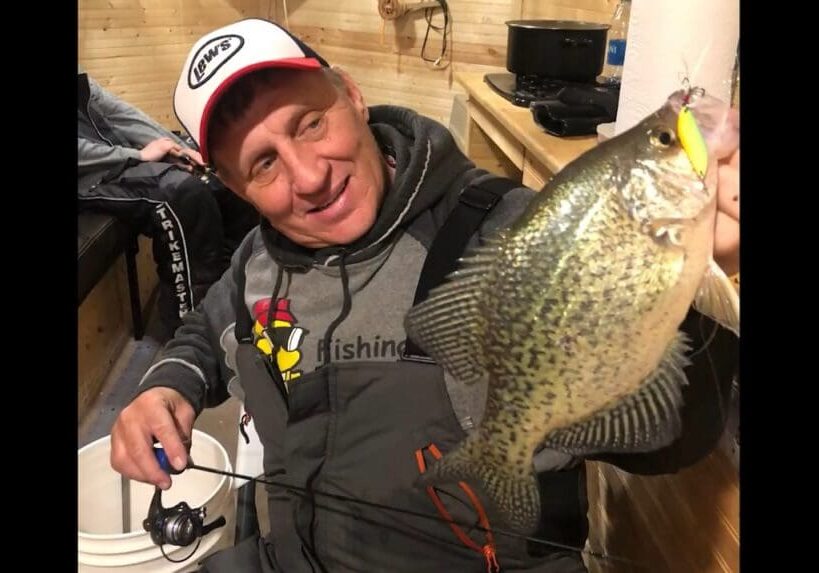 A smiling man in fishing gear, possibly Mike Frisch, holds up a large crappie fish indoors, sitting on a white bucket with a fishing rod in his other hand.