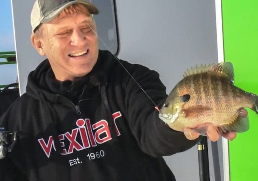 A smiling man named Mike in a cap and black "Vexilar" hoodie holds up a large sunfish by the mouth with a fishing rod in one hand, standing in front of a gray and green background.