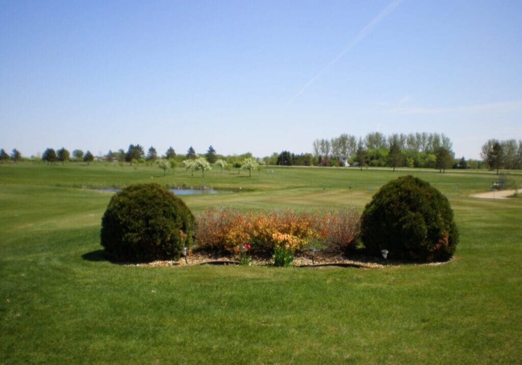 A landscaped flower bed with bushes and colorful flowers sits on the green at Nordic Trails Golf Course under a clear blue sky, with trees and a small pond in the background.