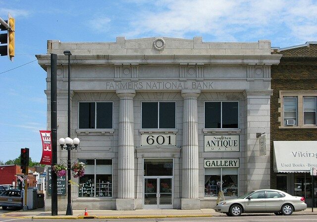 A historic stone building with columns labeled "Farmer's National Bank" houses Now & Then Antiques and a gallery at 601. A car is parked in front, with other businesses visible on either side.