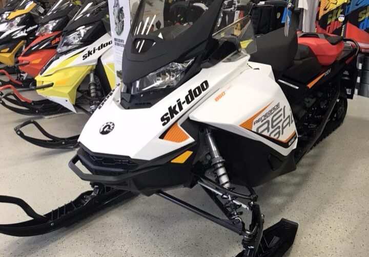 A white Ski-Doo snowmobile is displayed in Ollie's Service showroom, surrounded by other snowmobiles and colorful jackets hanging on the wall. A "Restroom" sign is visible in the background.