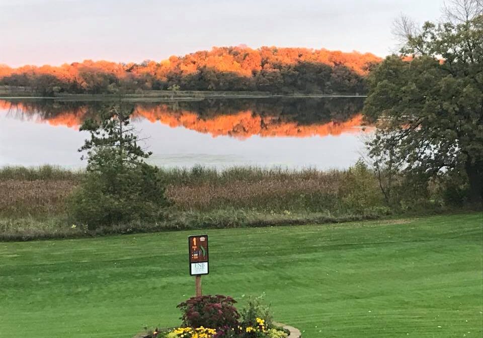 A grassy lawn at Osakis Country Club with a flower bed in the foreground overlooks a calm lake. Autumn trees with orange foliage are reflected in the water under an overcast sky, while a small sign stands near the flower bed.