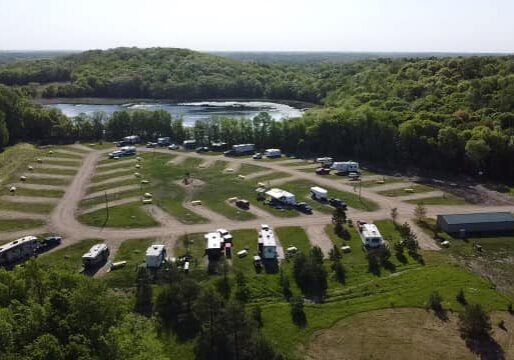 Aerial view of Andes RV Park campground with multiple parked RVs and trailers, winding gravel roads, scattered trees, a building, and a pond surrounded by dense green forest in the background.