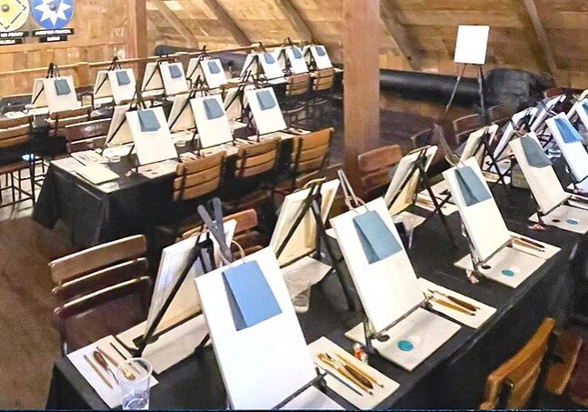 A room in the Palette District with rows of tables set up for a painting class, each featuring a canvas on an easel, paintbrushes, palettes, and cups. Wooden walls and a sloped ceiling complete the creative setting.