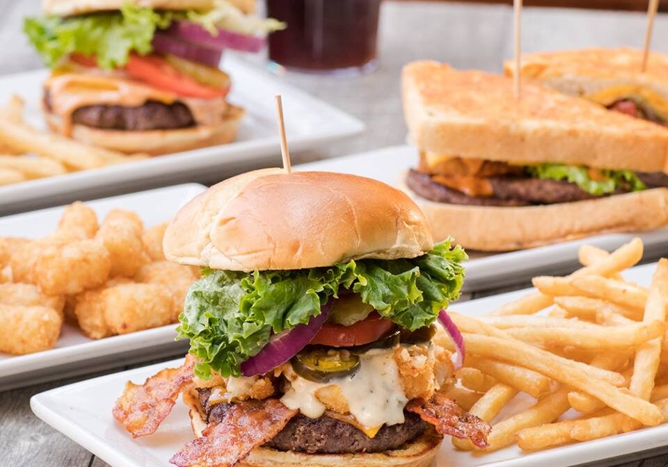 A close-up of a cheeseburger with bacon, lettuce, tomato, and onion, served with French fries at Perkins Restaurant. In the background are two more burgers and tater tots, all on white plates atop a wooden table.