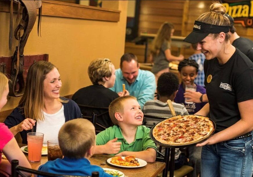 A smiling server presents a pizza to a happy family at Pizza Ranch, as children and adults enjoy their meal and conversation in a casual, welcoming atmosphere.