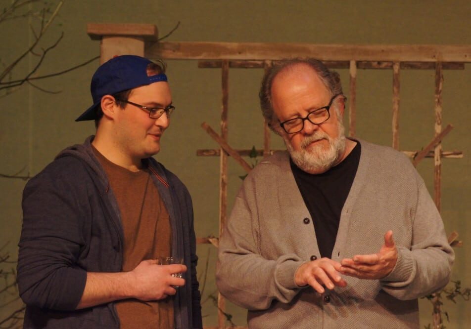 Two men stand together indoors. The older man with glasses and a gray sweater gestures as he talks, while the younger man in a blue cap and hoodie holds a glass and listens—capturing a scene from the award-winning show "Proof" at AAAA theatre.
