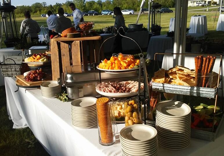 A Raaper's buffet table outdoors displays stacked plates, pretzel rods, assorted cheeses, crackers, fruit, and cold cuts. Guests serve themselves in the background under a canopy on a sunny day.