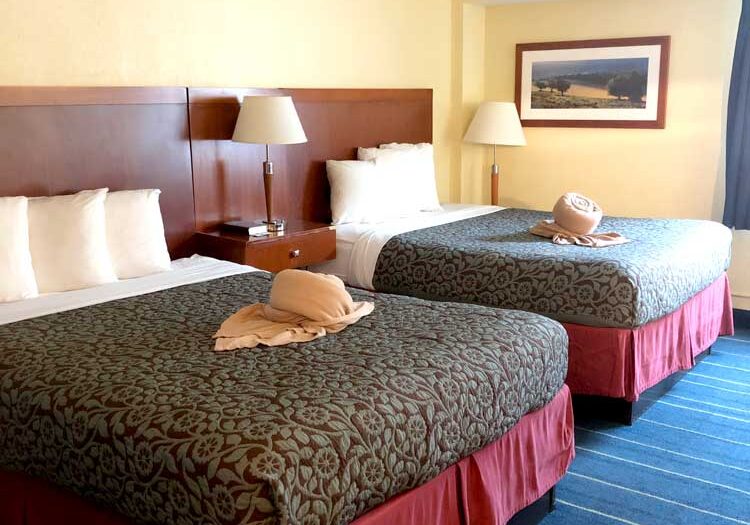 Experience comfort at Relax Inn, one of the top hotels in Alexandria Minnesota. This hotel room features two double beds with patterned bedspreads, towel art, white pillows, bedside tables with lamps, framed artwork, and striped blue carpet.