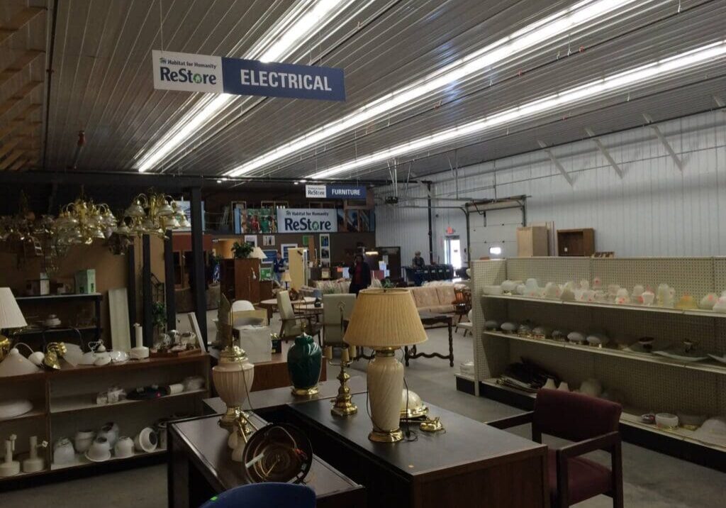 A Habitat for Humanity ReStore thrift store interior features shelves with lamps, furniture, and various home goods. Overhead signs label sections as "ELECTRICAL" and "FURNITURE." The space has metal walls, a high ceiling, and fluorescent lighting.