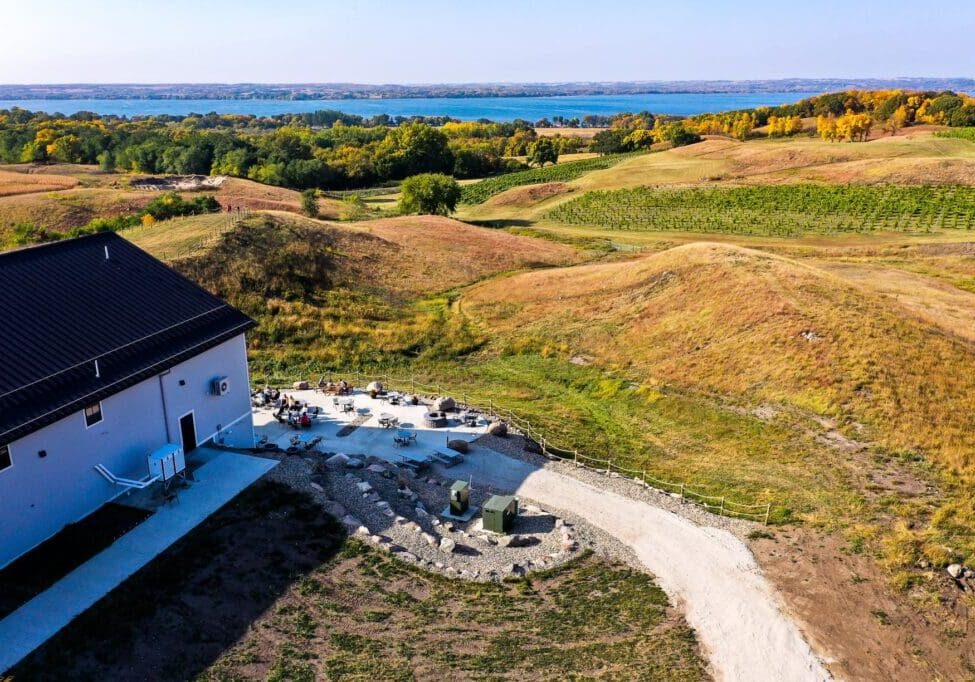 A white building sits beside a patio with tables and people at Rolling Forks Vineyards, surrounded by grassy hills and a gravel path, overlooking a river and tree-lined landscape under a clear blue sky.