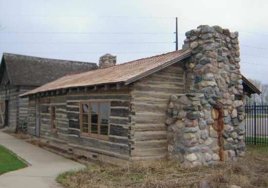 A rustic log cabin with a large stone chimney stands beside a paved sidewalk near the Runestone Museum. The cabin features a wooden exterior, shingled roof, and large windows, with dry grass along its foundation.