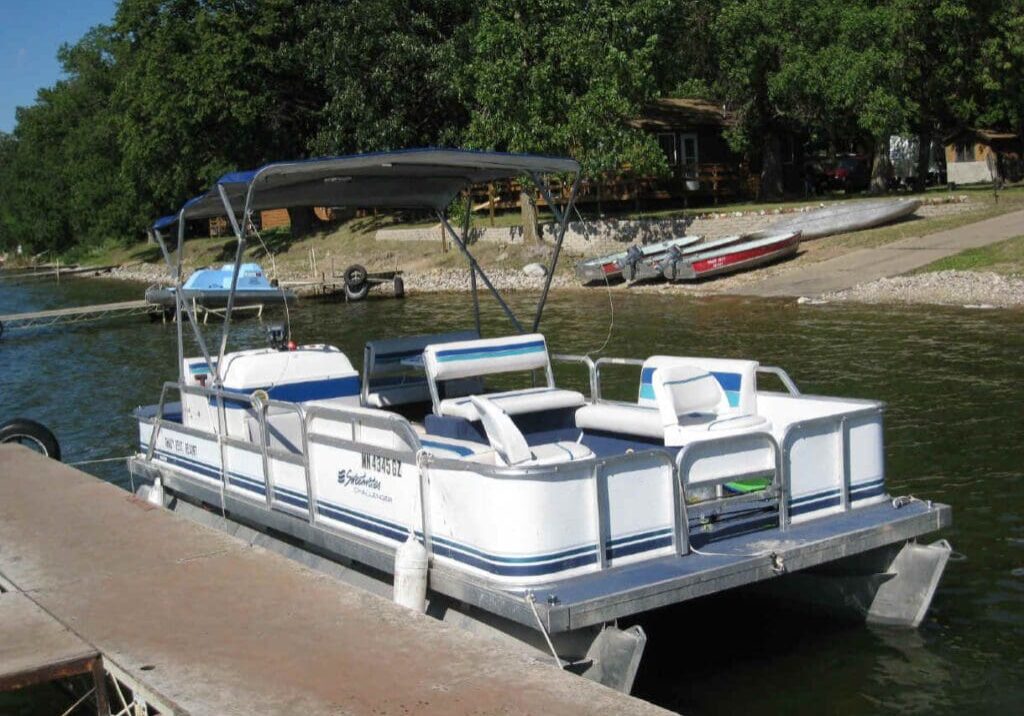 A white pontoon boat with blue stripes is docked at a lakeside pier at Shady Rest Resort & Campground. Trees, a house, and small boats dot the grassy, sloped shoreline in the background. The boat features blue and white seating and a canopy.