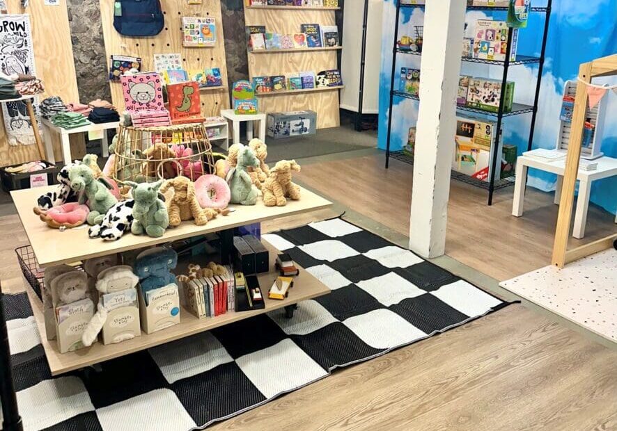 A cozy children’s shop, Lulie Boutique offers plush toys, books, and games on charming shelves and tables. With wood flooring, a checkered rug, and colorful decor, the space creates a playful and inviting atmosphere.