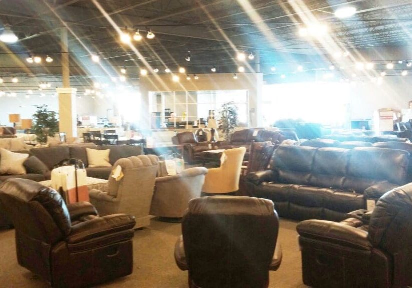 A large, brightly lit Slumberland Furniture showroom displays various sofas, armchairs, and recliners arranged in groups. The space has a high ceiling with exposed beams and many hanging lights. Sunlight streams in from large windows.