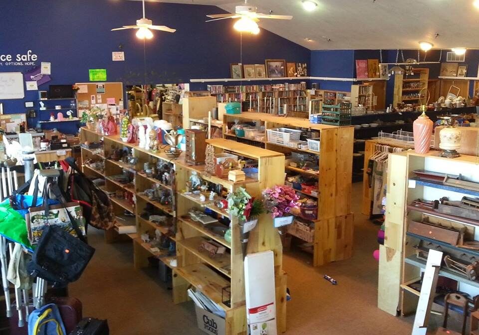 Someplace Safe Thrift Shop is a spacious store with wooden shelves displaying vases, knick-knacks, bags, books, and suitcases. It features beige carpet, a blue accent wall, ceiling fans, and fluorescent lights for a welcoming atmosphere.