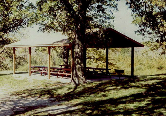 A wooden picnic shelter with a roof stands in Spruce Hill Park’s grassy area, shaded by large trees, with several picnic tables underneath and sunlight filtering through the leaves.