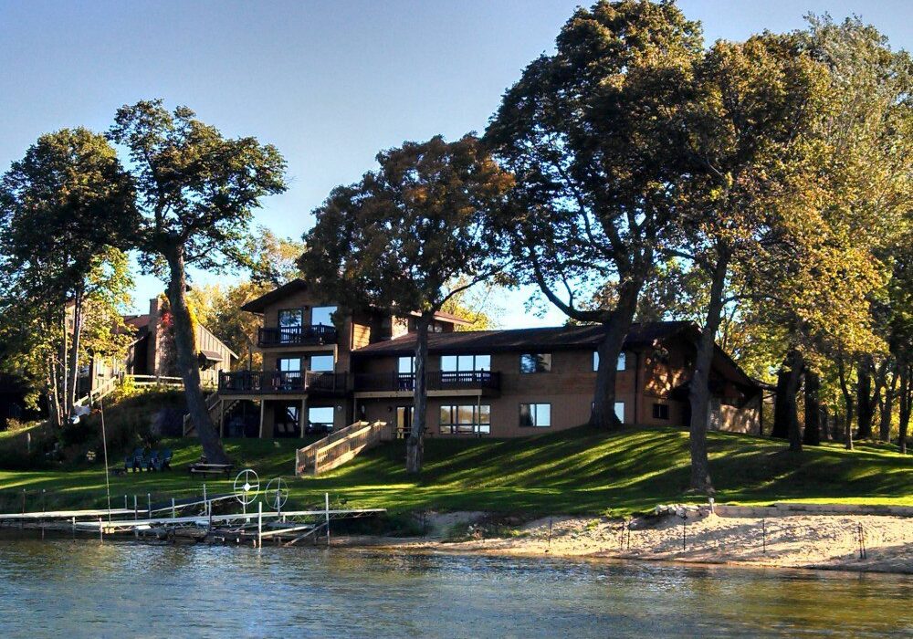 A large lakeside house named Sugarbush Lodge features multiple balconies and big windows, surrounded by tall trees and grassy lawn, with a sandy shore and a small dock leading into the water in the foreground.