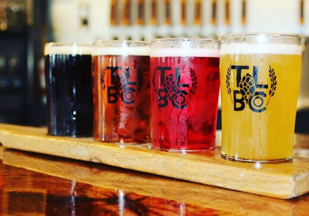 Four glasses of beer in varying hues, from dark brown to light yellow, are arranged on a wooden paddle atop a polished bar counter at Thousand Lakes Brewing, with beer taps softly blurred in the background.