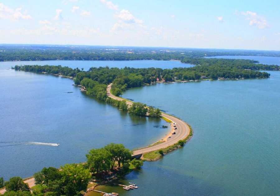 A winding road bordered by trees stretches across a narrow strip of land between two large blue lakes, with boats on the water and a bright, clear sky—one of the region’s must-see attractions.