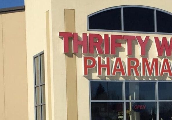 The front of a Thrifty White Drug Pharmacy building with large red letters spelling the store name above the entrance. A lit "OPEN" sign in the window indicates the store is open for customers.