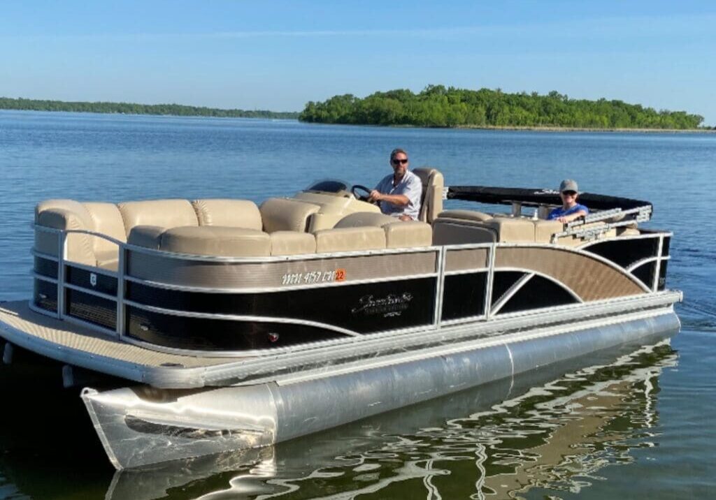 A man and a child ride a beige and black pontoon boat on calm water, with a small green island and clear blue sky in the background—another perfect outing from Toon Tastic Pontoon Rentals.