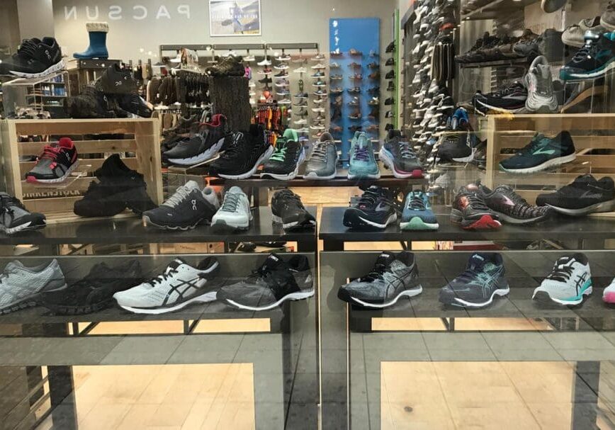 A store display for Tradehome Shoes showcases a variety of athletic shoes in different styles and colors, arranged on shelves and platforms behind a glass window. The background features even more shoes and store fixtures.