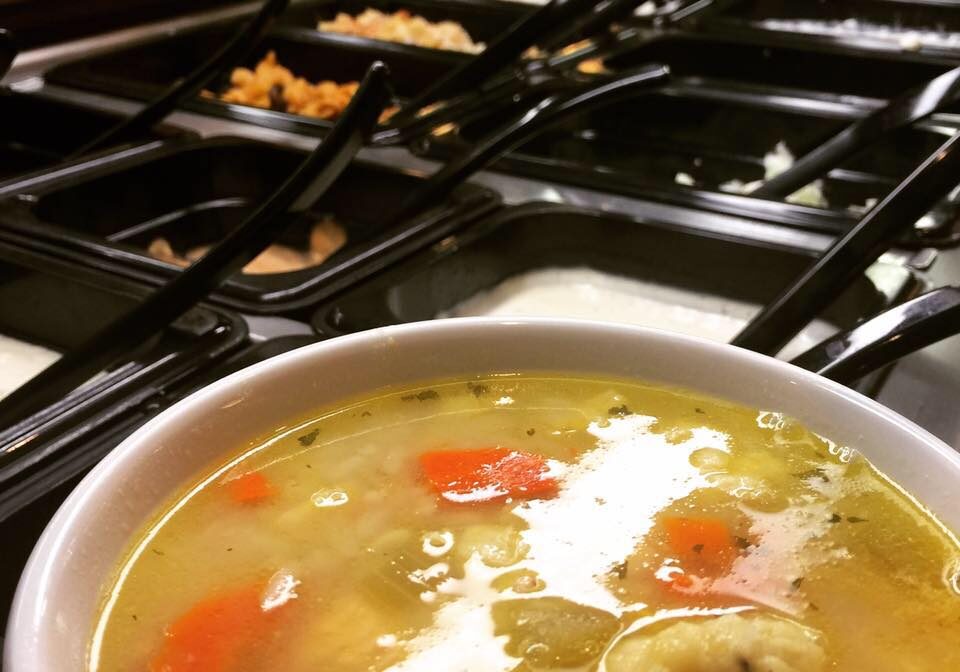 A close-up of a bowl of vegetable soup with carrots and potatoes, served at Traveler's Inn Cafe, sits in front of a self-serve salad bar featuring various toppings and dressings in black containers.