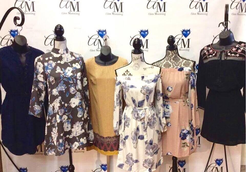 Six mannequins display a variety of women's dresses in floral and solid patterns, arranged in front of a white backdrop with blue and black "CM Glen Manning" logos, embodying the latest trend and couture styles.