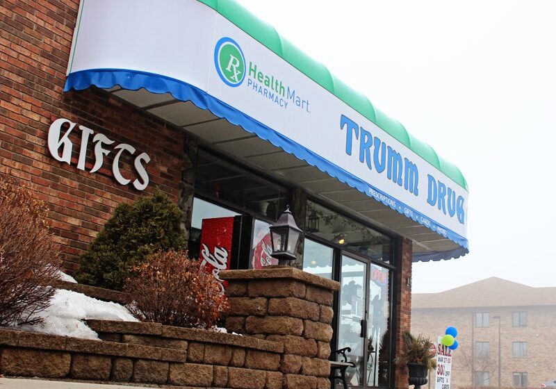 A brick building with a blue and green awning that reads "TRUMM DRUG" and "Health Mart Pharmacy." The Trumm Drug entrance displays "GIFTS," with snow on the ground and shrubs near the door.