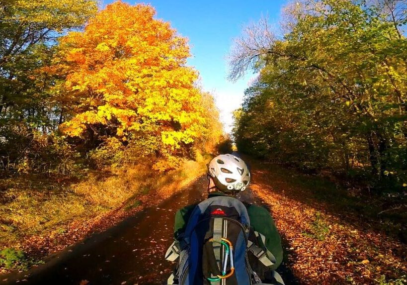 A person biking across Minnesota, wearing a helmet and backpack, cycles along a tree-lined path in autumn, with vibrant yellow and orange foliage under a clear blue sky.