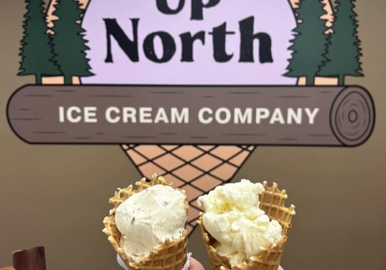 Two hands hold waffle cones topped with creamy scoops, lifted in front of the Up North Ice Cream Company sign, which features a rustic log and pine tree design.