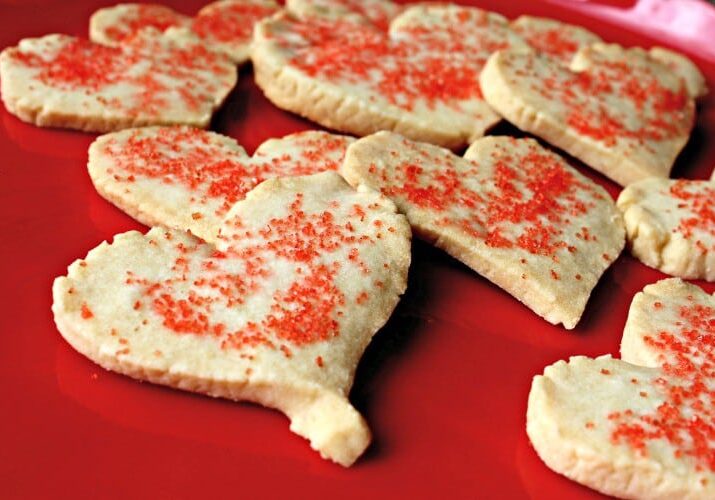 Heart-shaped sugar cookies topped with red sprinkles are arranged on a bright red surface, adding a touch of fun and romance perfect for Valentine’s Weekend.