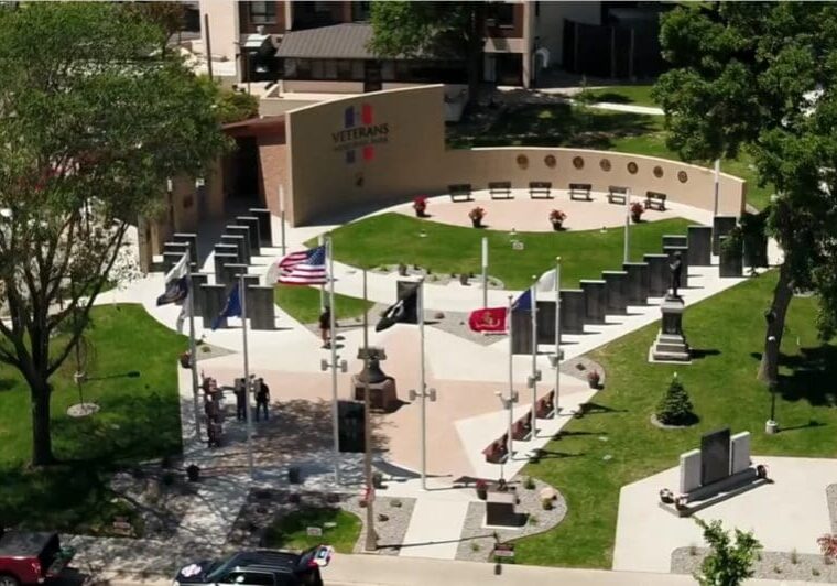 Aerial view of Veterans Memorial Park featuring a curved wall with emblems, multiple flagpoles with flags, a statue, paved walkways, green grass, benches, and trees surrounding the area.