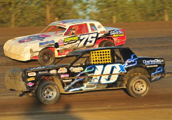 Two dirt track race cars, one white with number 75 and one black with number 10X, speed side by side at Viking Speedway, with trees blurred in the background.