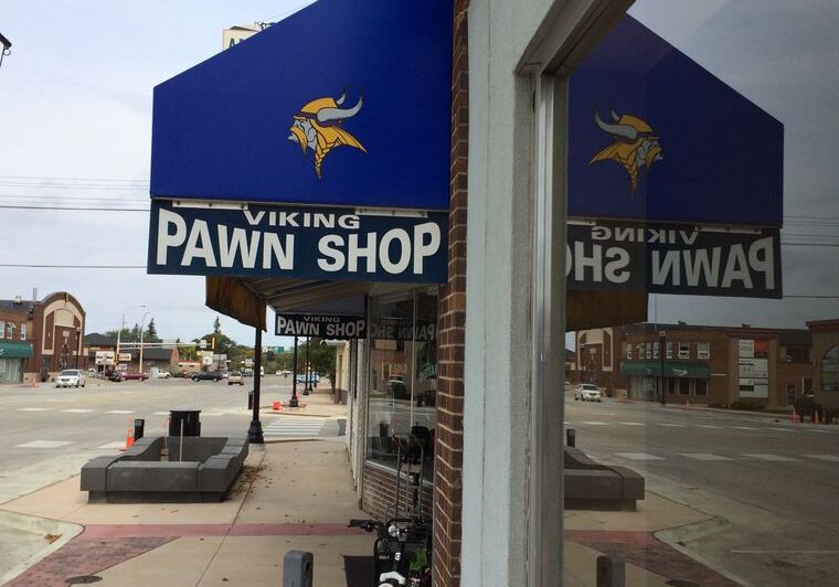 A blue awning with a Viking logo and the words "Viking Pawn Shop" covers the quiet sidewalk outside the Viking Pawn Shop, its large window reflecting the storefront and nearly empty street.