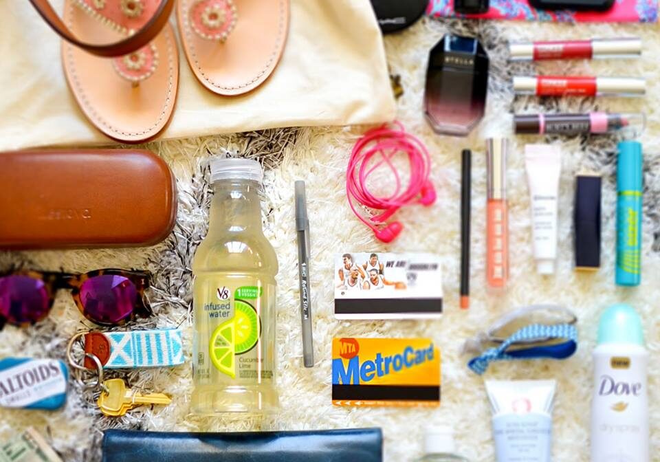 A variety of items laid out, including sandals, sunglasses, cash, a wallet, keys, a MetroCard, lipstick from Walgreens, skincare products, headphones, a drink, mints, and a phone, all arranged on a flat surface.