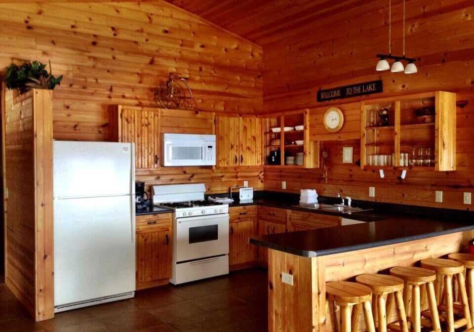 A cozy kitchen at Woodland Resort with wooden walls, cabinets, and ceiling features a white refrigerator, stove, and microwave. There’s a counter with four wooden stools, shelves with dishes and glasses, and a “Welcome to the Lake” sign above.