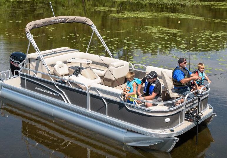 A family of four, all wearing life jackets, enjoys fishing from a pontoon boat on a calm lake surrounded by greenery and lily pads. Relaxing on the deck, they make the most of their day with Rentals at Woodland Resort.