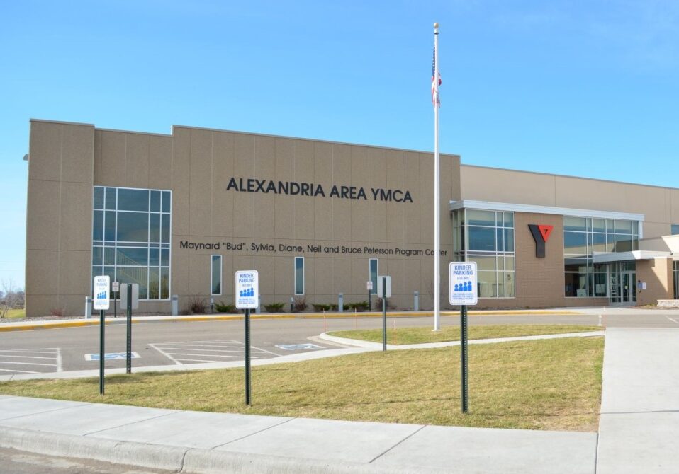 Exterior view of the Alexandria YMCA building with large windows, a tan facade, and signs indicating accessible parking spaces in front, all beneath a clear blue sky.