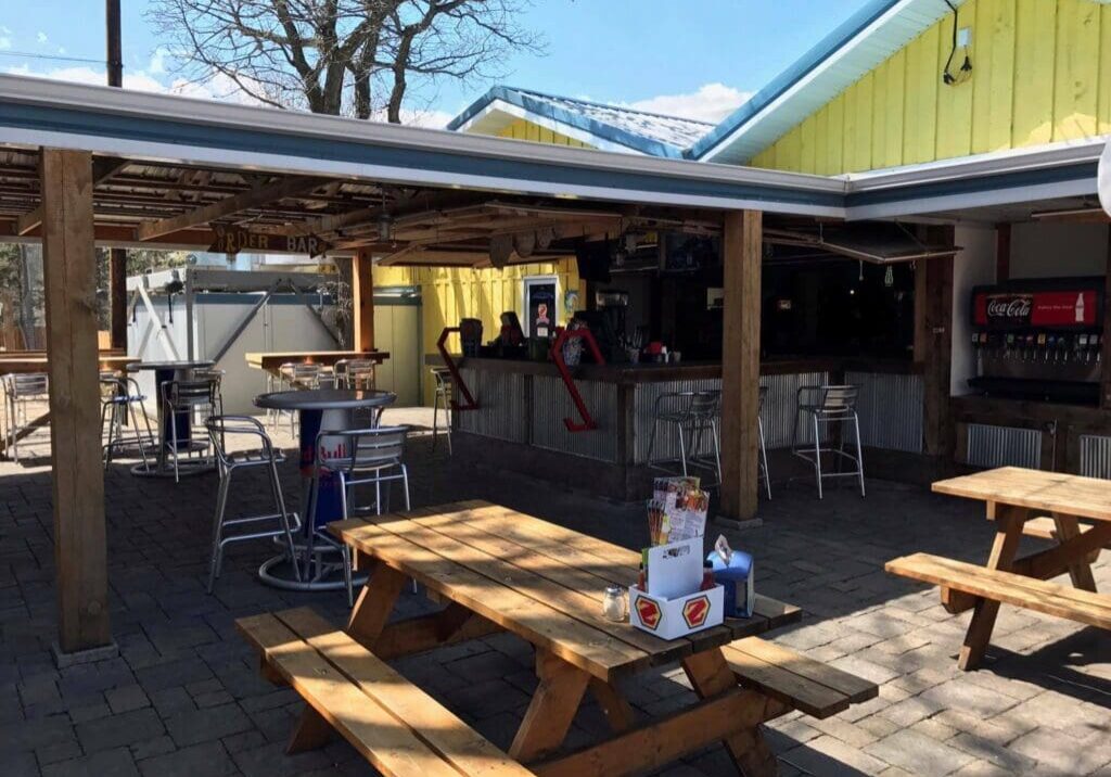 Outdoor bar and seating area with wooden picnic tables, metal stools, a Zorbaz Red Bull table, and a Coca-Cola soda dispenser. The bar sits under a wooden roof with bright yellow walls in the background on a sunny day.